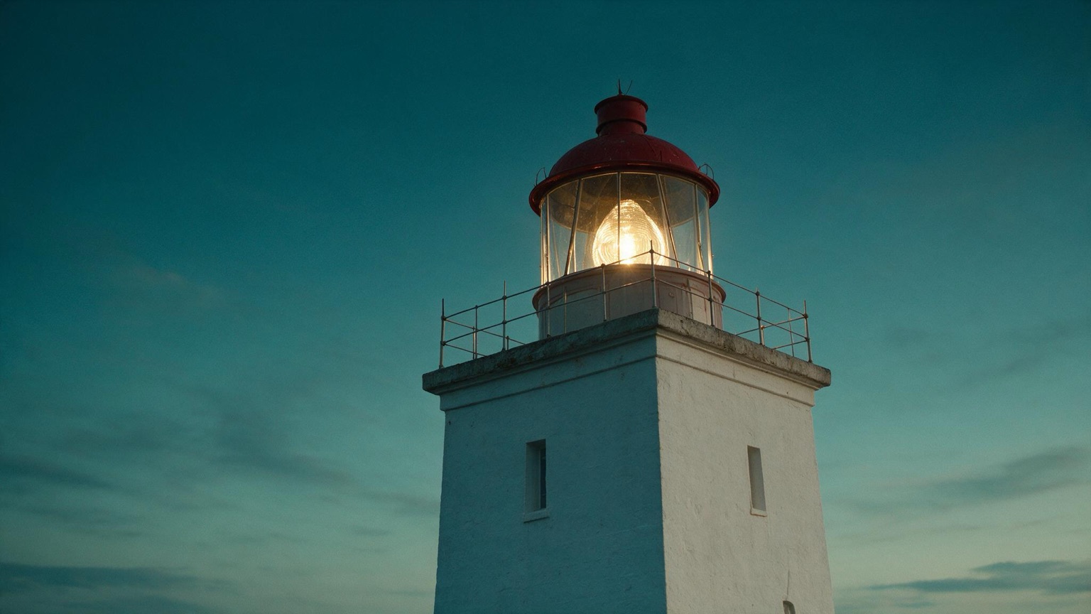 Close view of the same lighthouse lamp room at dusk, the beacon lit warm behind its glass against a teal sky